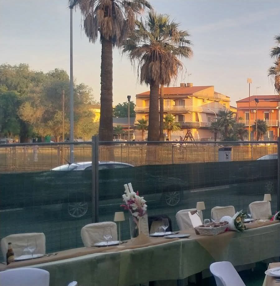 Tavolata all'aperto allestita dal catering al tramonto: tovaglie verdi e color sabbia, fiori bianchi e rosa, lampade da tavolo e sedie coperte, con le palme sullo sfondo