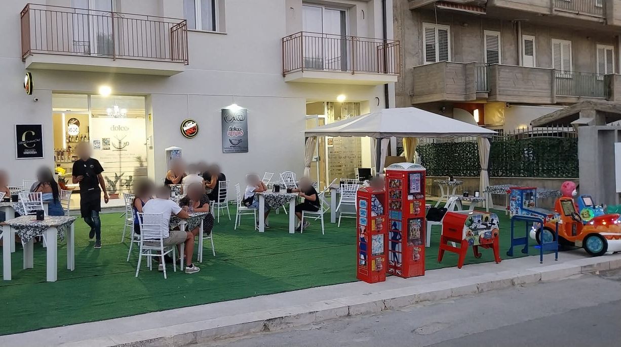 Il dehors di Dolce Amaro su Via Tarantelli: tavolini bianchi sul prato sintetico, il gazebo e i clienti seduti all'aperto al tramonto